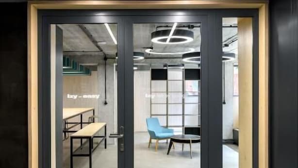 A modern office space with a glass partition, a blue chair, and a wooden ceiling with linear lighting. - IZY STAY Hostel Bratislava (Bratislava)