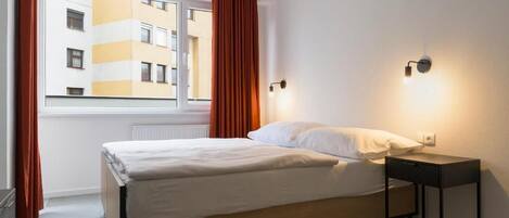 A bedroom with a bed, a nightstand, a window with curtains, and a wall-mounted light fixture.