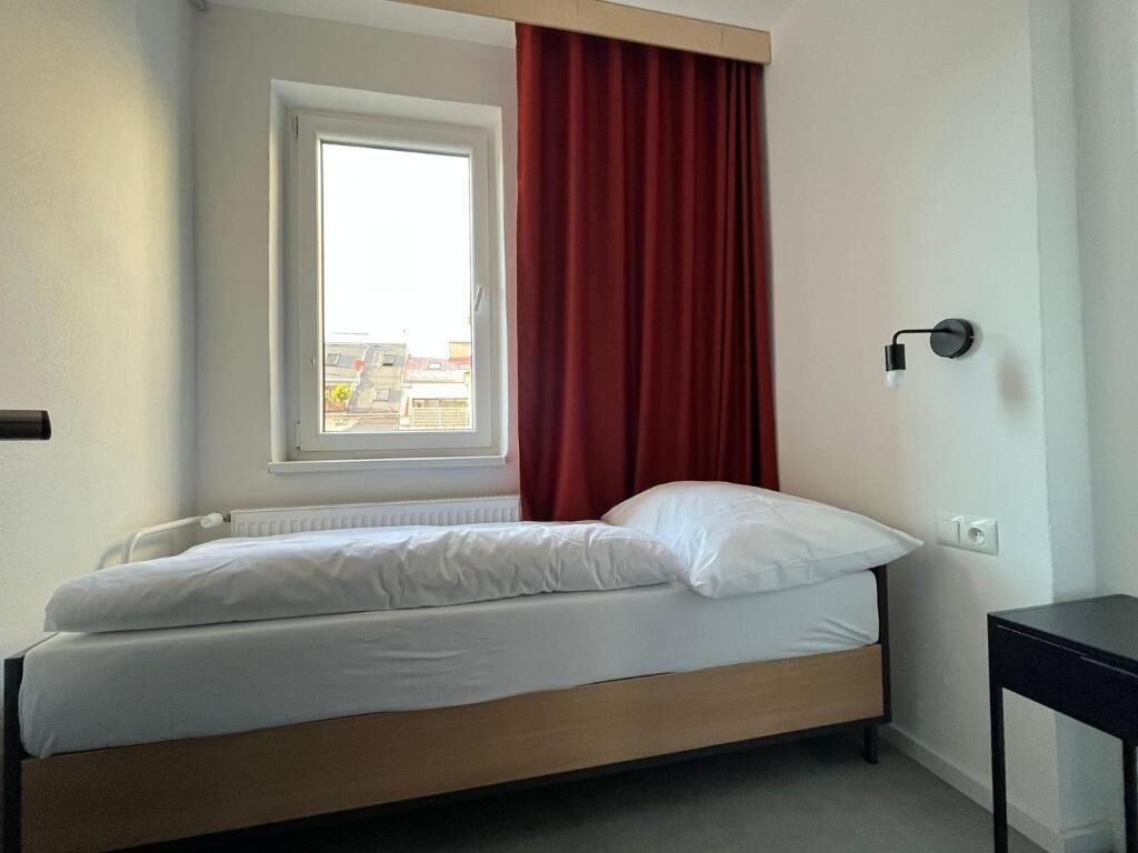A single bed with white bedding in a room with a window and red curtains.