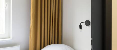 A single bed with a wooden frame, a white mattress, and a white pillow. There is a window with a white frame and a yellow pendant light above the bed. A brown curtain is partially drawn to the side.