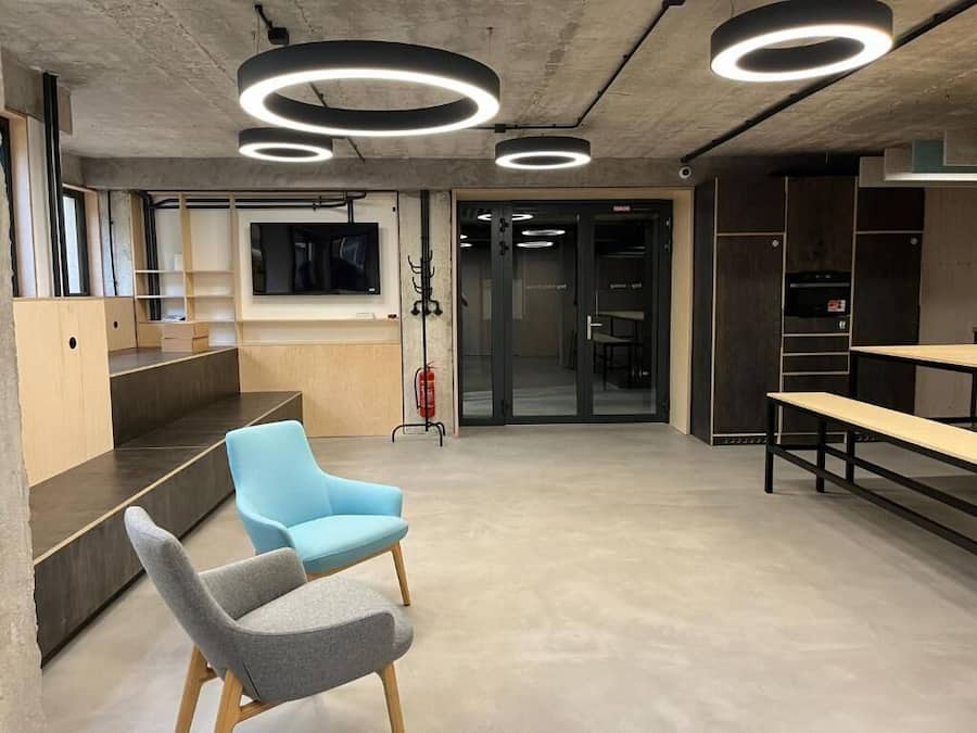 Een moderne kantoorruimte met een betonnen plafond, ronde lampen, een grijs- en blauwe stoel, een houten bank en een brandblusser.