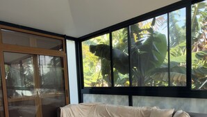 A modern living room with a grey sofa, a large window with a view of tropical plants, and a sliding glass door. - 3-bedroom house in Bananeiras in the center of Funchal (Funchal)