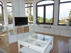 Une pièce spacieuse avec de grandes fenêtres, un téléviseur posé sur un meuble blanc et une table à plateau en verre.