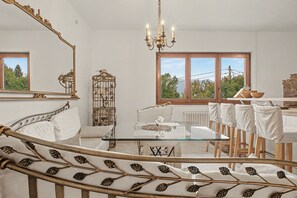 Ein Zimmer mit einem Esstisch mit Glasplatte, weiß gepolsterten Stühlen, einem weißen Sofa und einem dekorativen Vogelkäfig aus Metall.