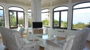A spacious living room with a glass coffee table, two white armchairs, a flat-screen TV, and a white cabinet. Large arched windows offer a view of greenery and a distant landscape. - Casa Paradiso in Porto Valtravaglia (Porto Valtravaglia)