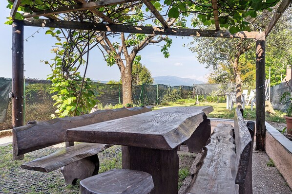 A wooden picnic table with benches under a pergola, surrounded by greenery and a view of distant mountains.