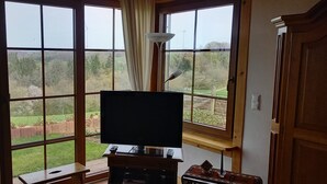 Een kamer met een groot raam, een televisie op een houten standaard, een klein tafeltje en een decoratief object op de vensterbank.