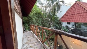 Un balcon doté d’un sol en pierre, d’une balustrade en bois et offrant une vue sur des palmiers et un bâtiment avec un toit rouge.