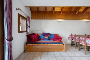A cozy bedroom with a wooden bed, a window with curtains, and a wooden table with a tablecloth. - Apartment 'Les Roches' with Mountain View (Saint-Colomban-des-Villards)