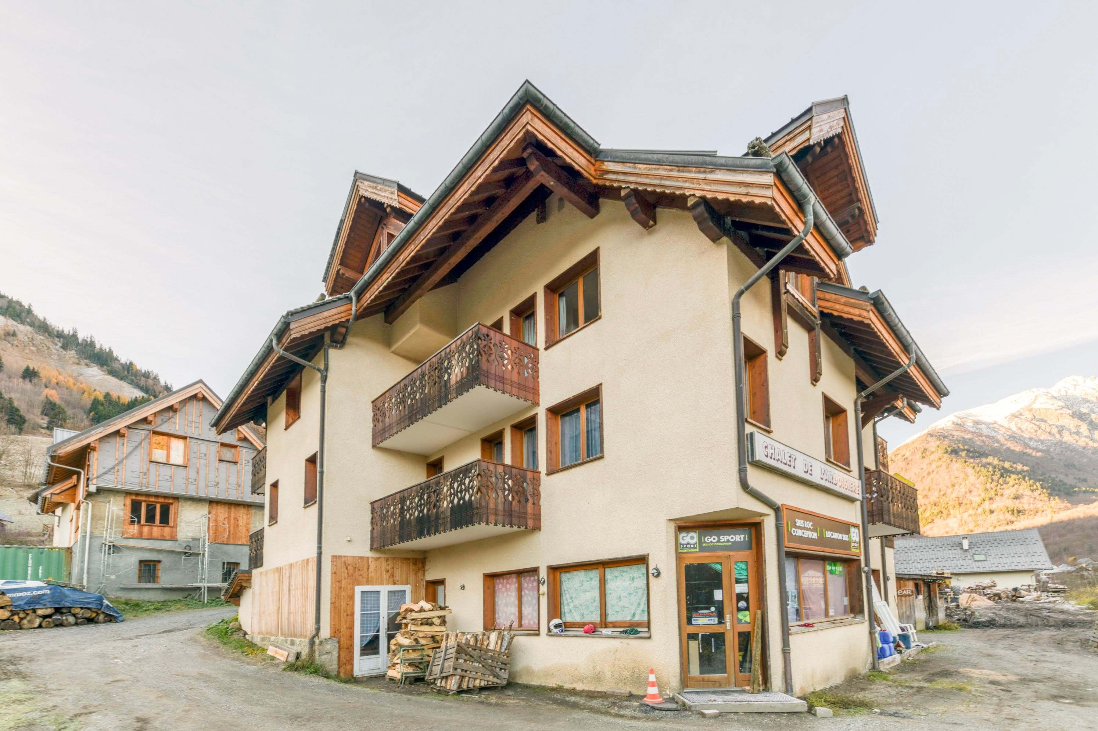Un immeuble à plusieurs étages doté de balcons, avec un commerce au rez-de-chaussée, et un paysage montagneux en arrière-plan.