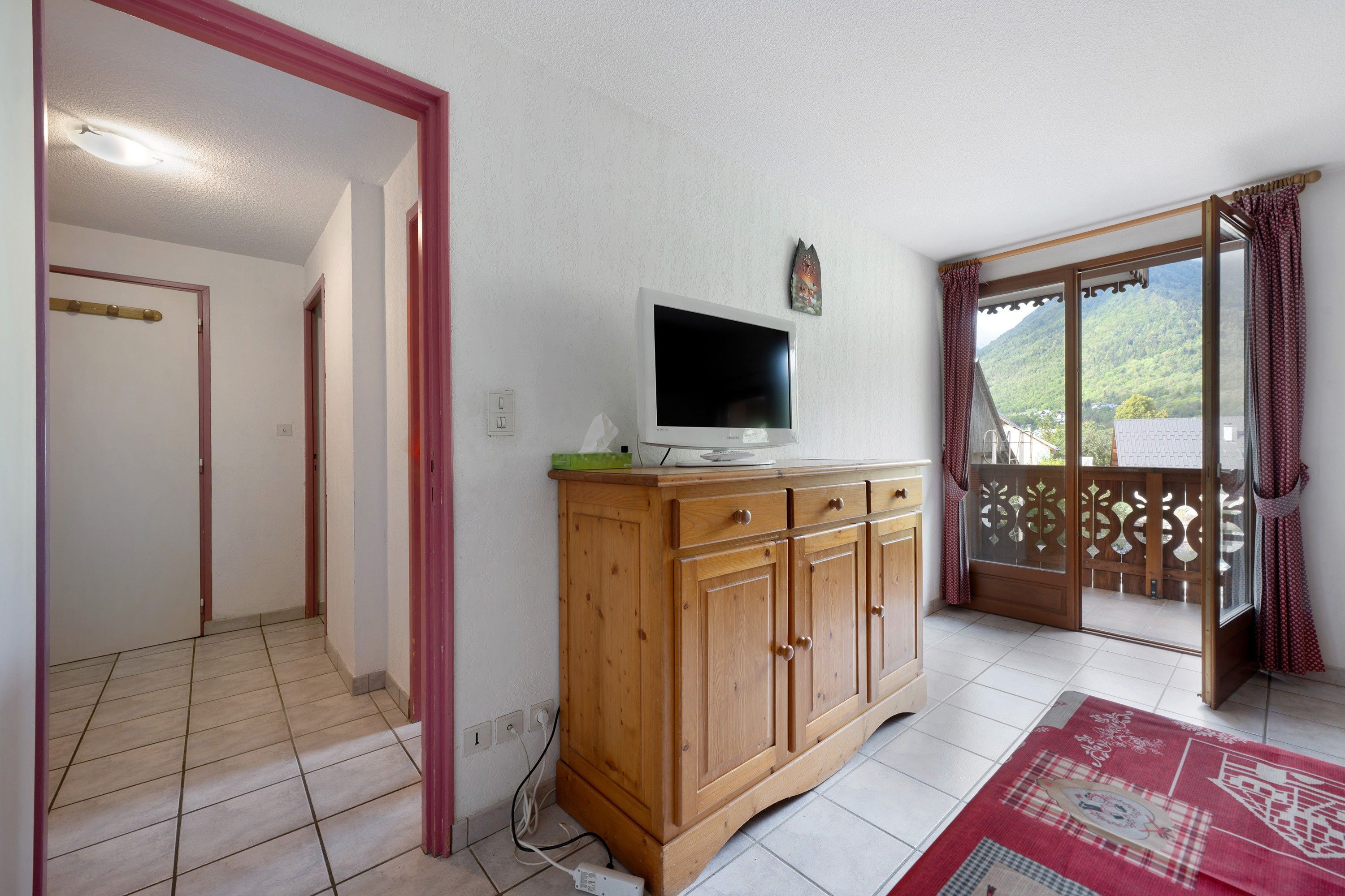 A room with a wooden cabinet, a television, and a balcony with a view of a mountainous landscape.