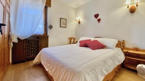 A bedroom with a large bed, wooden headboard, a bedside table, a nightstand, a window with a curtain, wall-mounted lamps, and decorative heart-shaped items. - Cozy 3 bedroom in amazing location (Cortina d'Ampezzo)