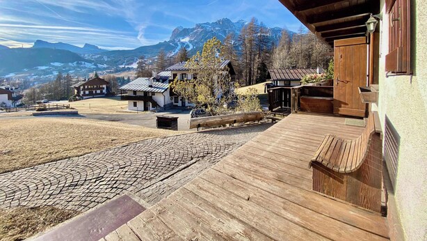 A wooden deck with a cobblestone path leading to a building with a traditional design, set against a backdrop of mountains and a clear sky. - Cozy 3 bedroom in amazing location (Cortina d'Ampezzo)