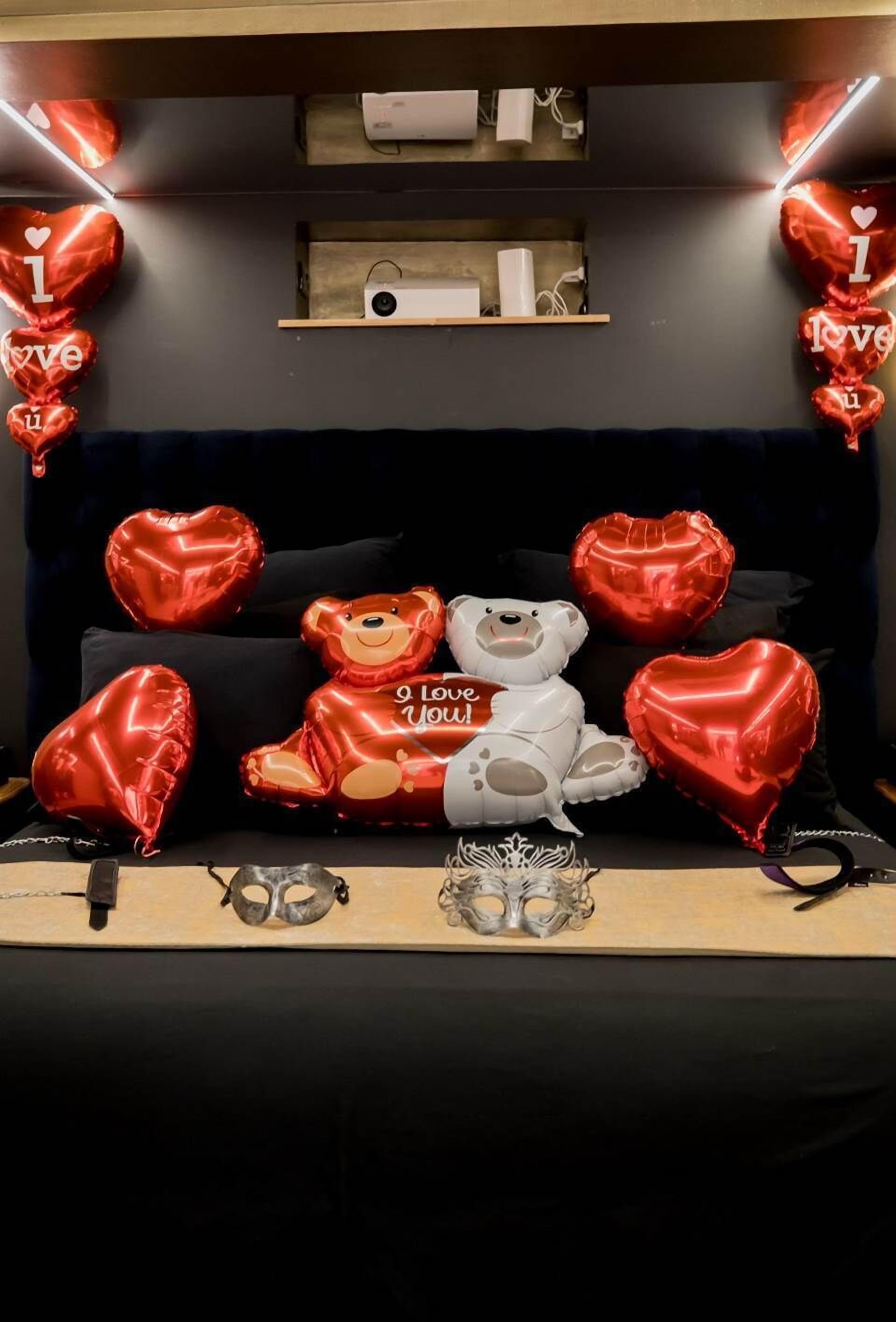 Una habitación con un sofá negro, dos osos de peluche, globos rojos con forma de corazón y un estante con un altavoz y otros objetos.
