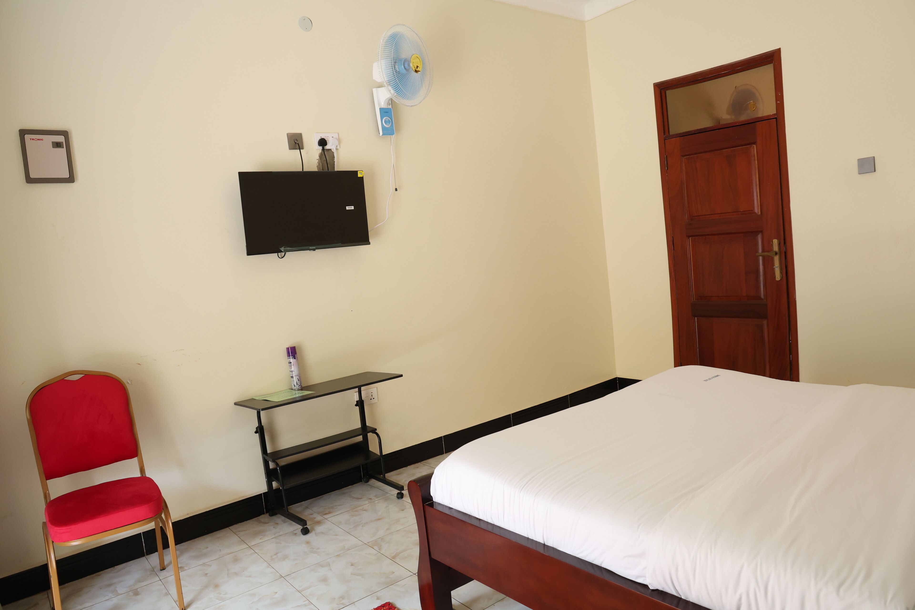 Una habitación con cama, escritorio, silla, ventilador de pared y puerta.