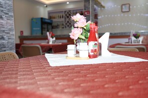 A table with a red tablecloth, a white napkin, a small vase with pink flowers, and a bottle of Shalal tomato sauce. - Simbego Cottages  (Lira)