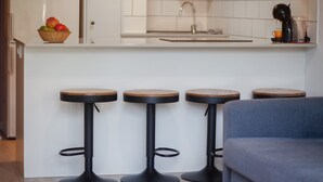 Eine Küchenzeile mit weißer Arbeitsplatte, Obstkorb, Kaffeemaschine und drei Hocker mit Holzbeschlägen.