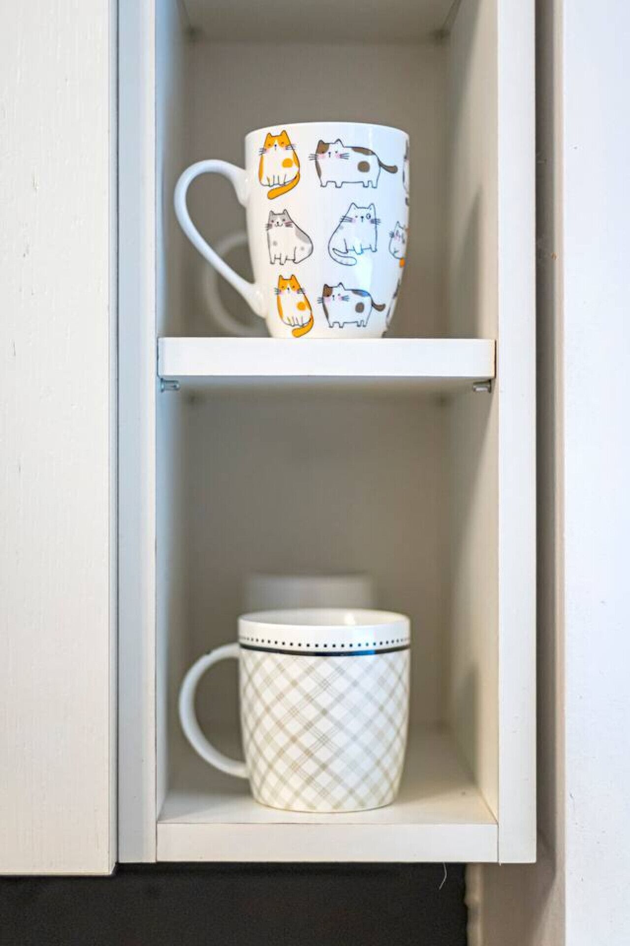 A white cabinet with two shelves; the top shelf holds a mug with cat illustrations, and the bottom shelf holds a mug with a plaid pattern.