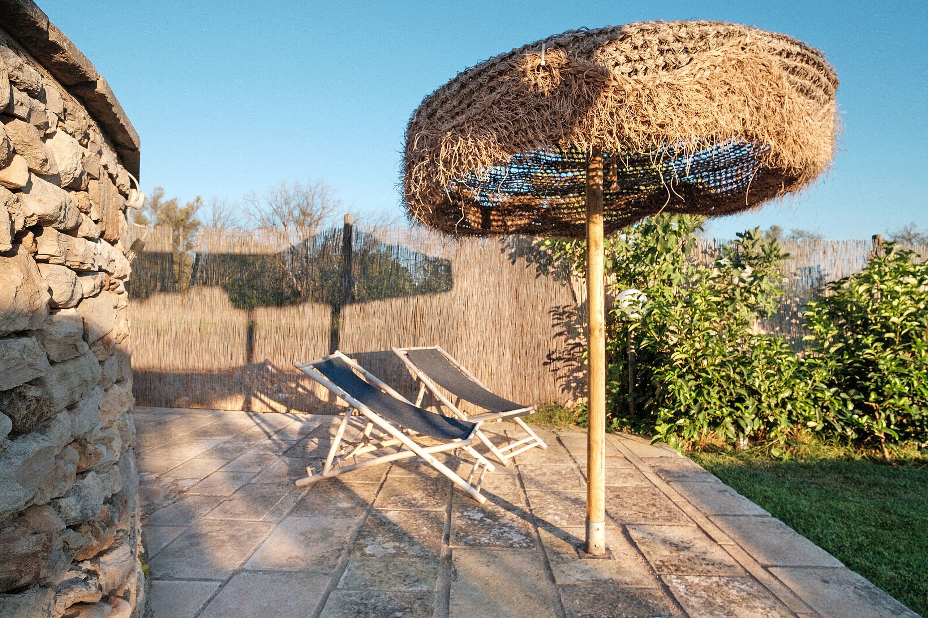 Un muro in pietra, un ombrellone di paglia e due poltrone su una terrazza.