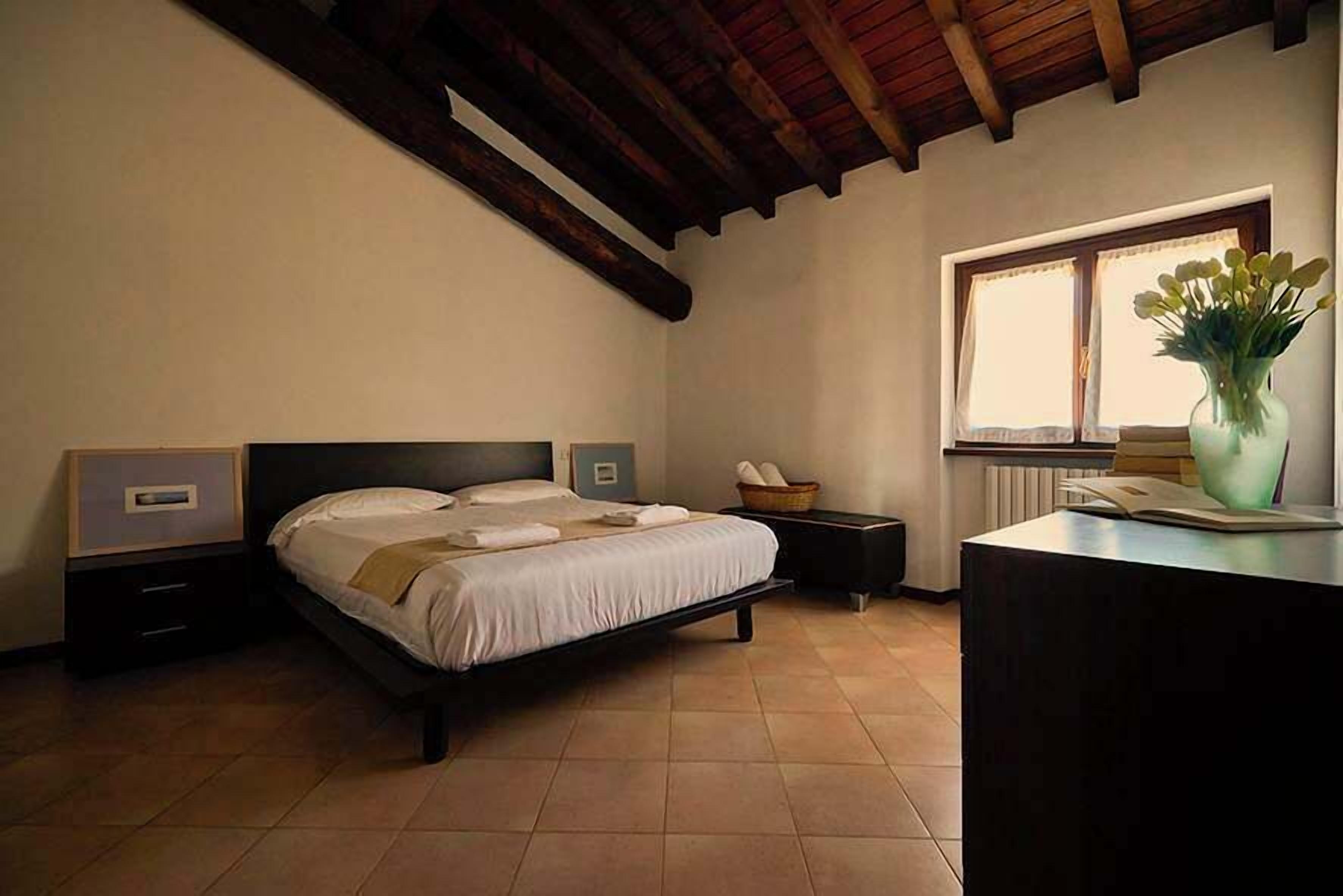 Une chambre à coucher avec un lit, des tables de chevet, une fenêtre avec des rideaux, un bureau et un vase avec des fleurs.