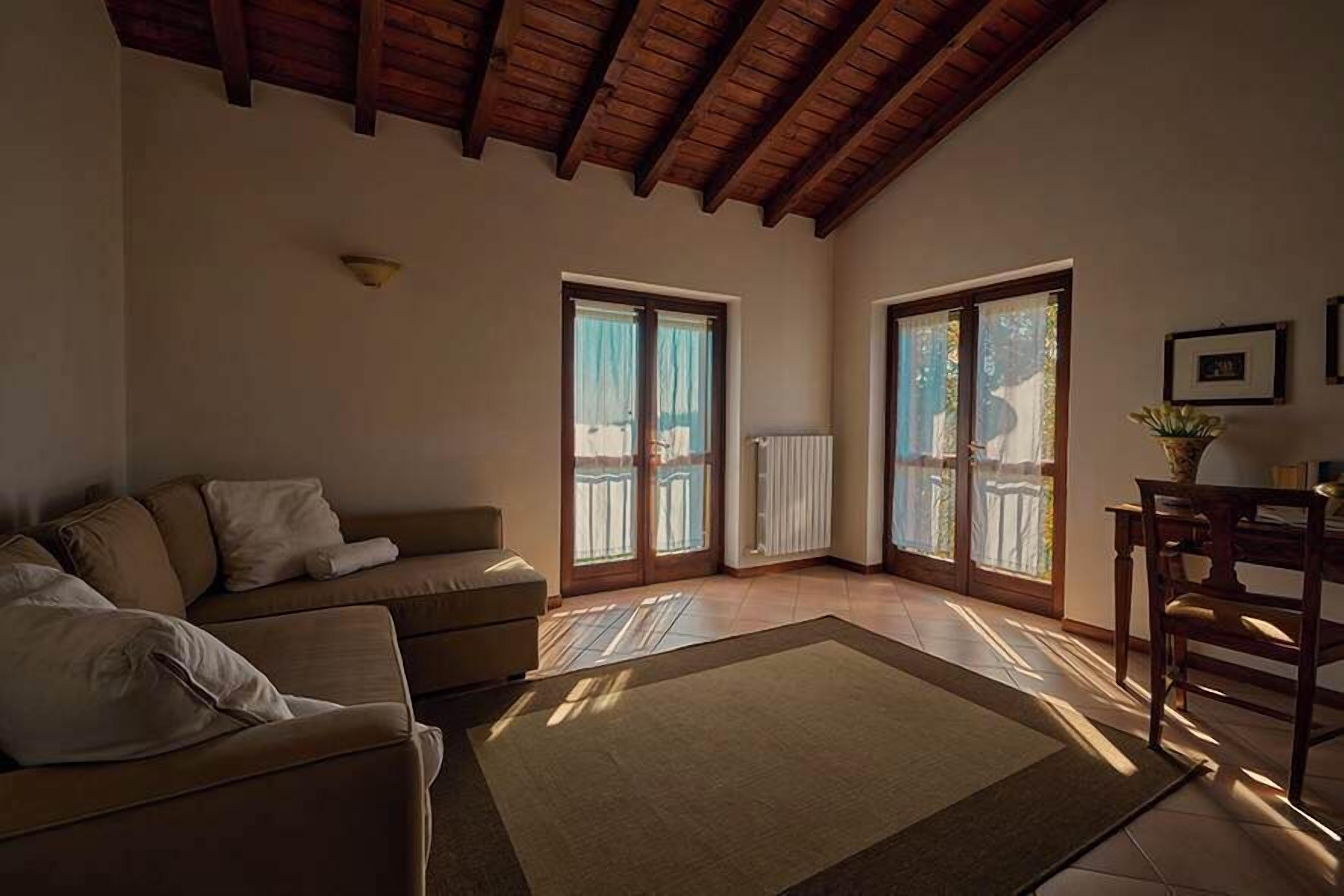 Una sala de estar con sofá, una mesa de centro y una mesa de comedor con sillas. Hay ventanas amplias con cortinas y un techo de madera.