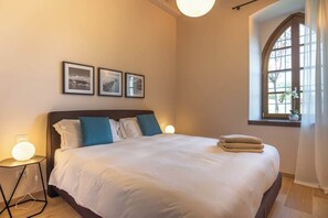 Ein ordentlich bezogenes Bett mit weißen Laken und blauen Kissen, ein Nachttisch mit Lampe sowie drei gerahmte Bilder an der Wand.