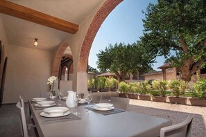 Un área de comedor al aire libre con una mesa larga dispuesta para una comida, rodeada de arcos de ladrillo y plantas en macetas.