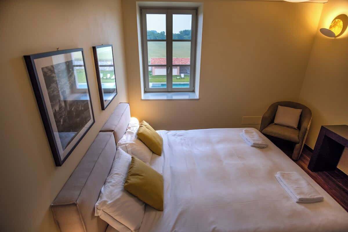 Una cama bien hecha con sábanas blancas y dos almohadas, una silla, una mesita y dos cuadros en la pared.