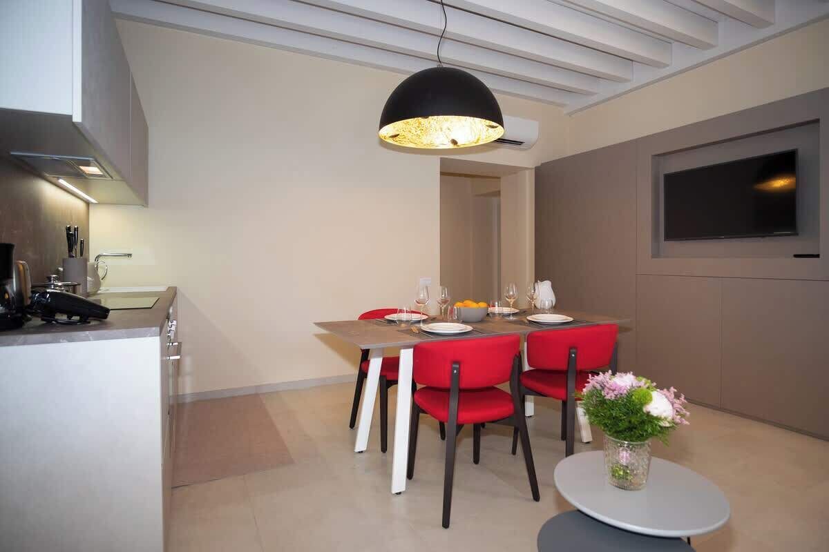 A modern dining area with a table set for four, red chairs, a small round table with a vase of flowers, and a ceiling light fixture.