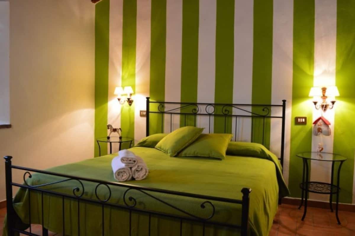 Una cama con una colcha verde, dos almohadas y una toalla enrollada. Hay dos lámparas de pared a cada lado de la cama.