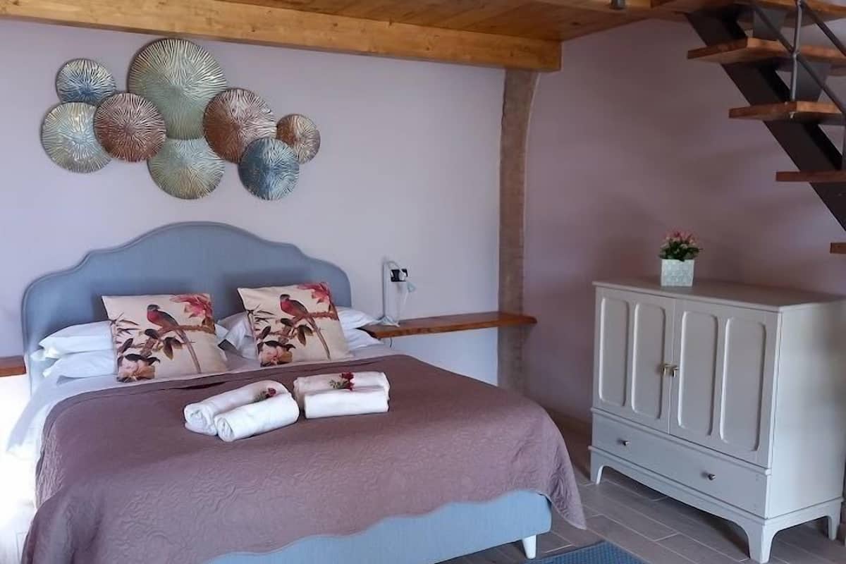 Un dormitorio con cama, almohadas, una mesita de noche, cómoda y una obra de arte en la pared.