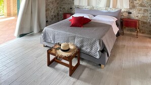 Une chambre avec des murs en pierre, un plafond en bois, un lit recouvert d’une couverture grise et agrémenté d’oreillers rouges, une petite table en bois sur laquelle repose un chapeau de paille, et une fenêtre ornée de rideaux blancs.