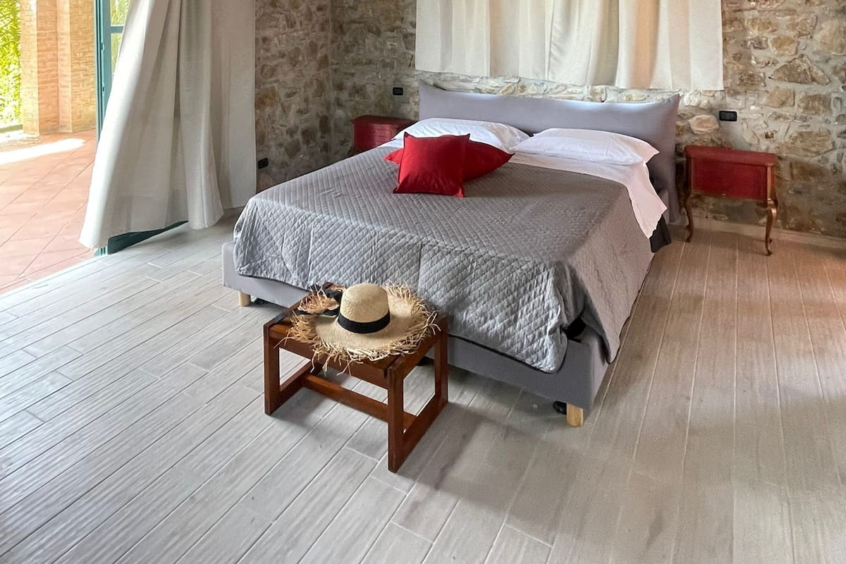 Un dormitorio con paredes de piedra, techo de madera, cama con cubrecama gris y almohadas rojas, una pequeña mesa de madera con sombrero de paja y una ventana con cortinas blancas.