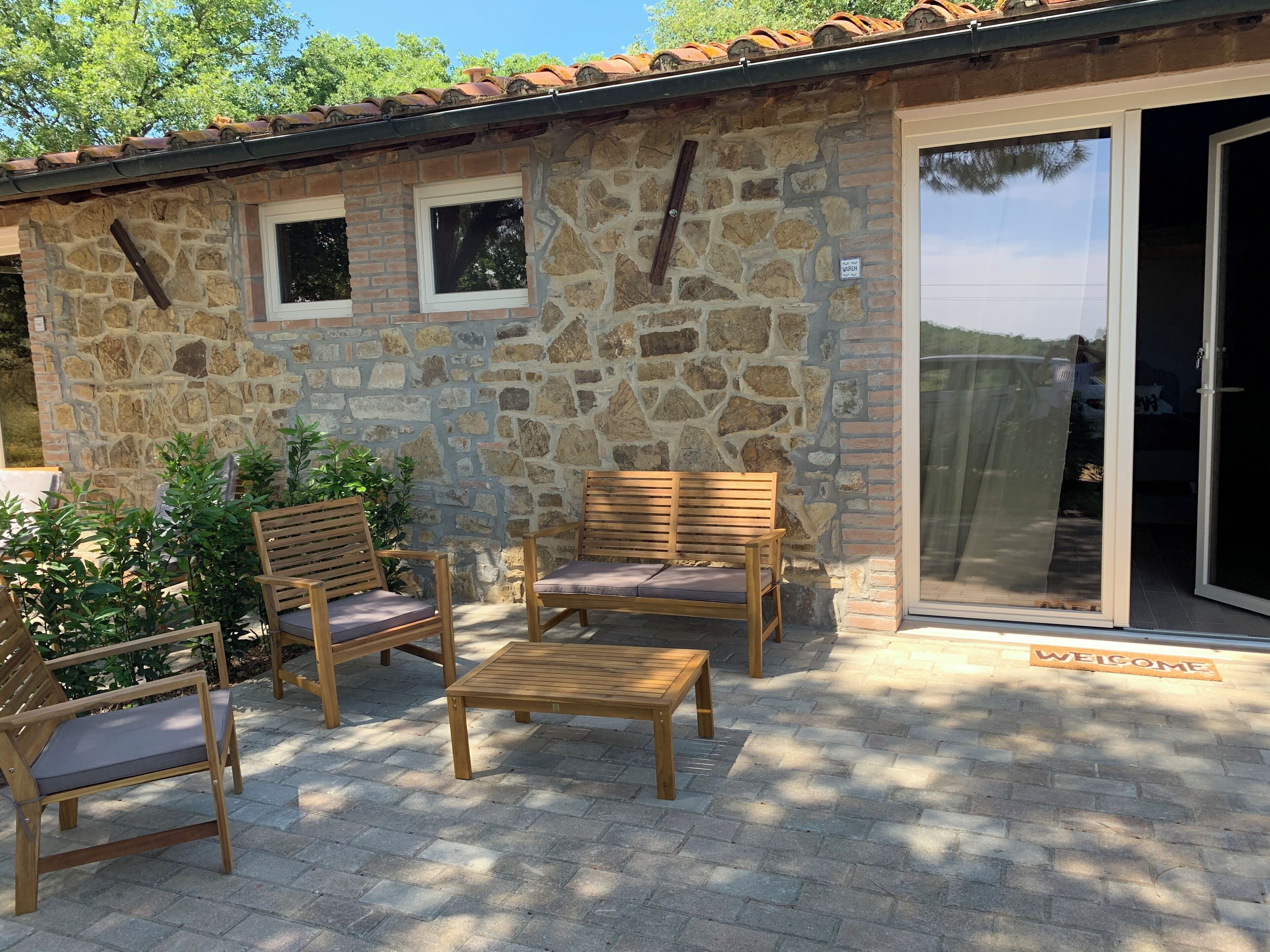 A stone building with a tiled roof, a wooden bench, and two wooden chairs with cushions.