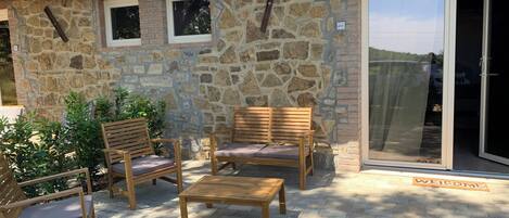 A stone building with a tiled roof, a wooden bench, and two wooden chairs with cushions.