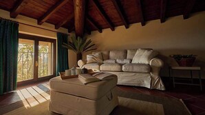 Ein Wohnzimmer mit einem beigefarbenen Sofa, dazu passendem Hocker, einem kleinen Tisch mit Pflanze und einem großen Fenster mit Vorhängen.