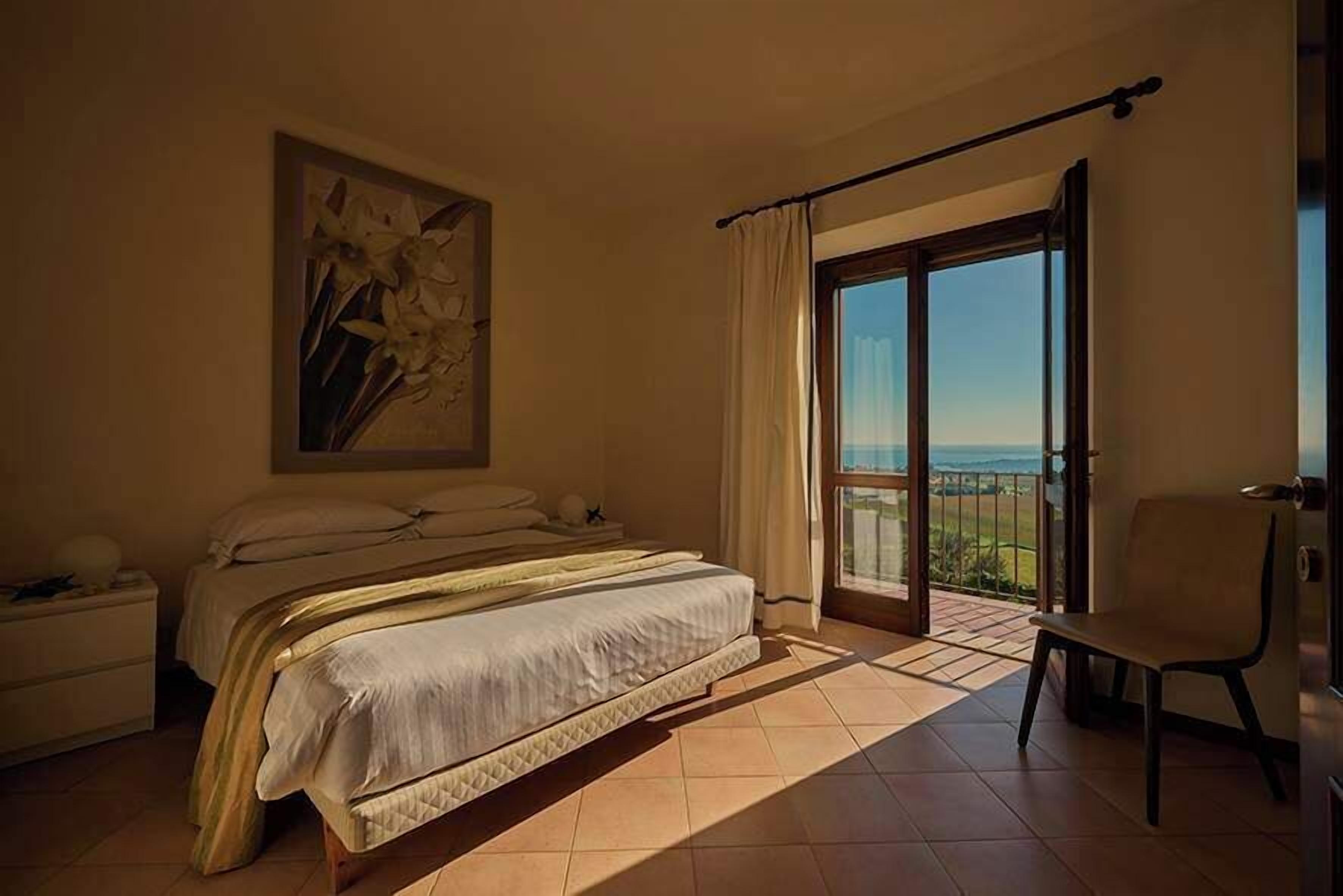 A bedroom with a bed, a nightstand, a chair, and a large window with a view.