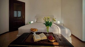 A neatly made bed with white linens, a bedside table with a book, a bowl of croissants, a vase of flowers, and a fruit bowl. - Villa Montecroce Apartment n.2 (Padenghe sul Garda)