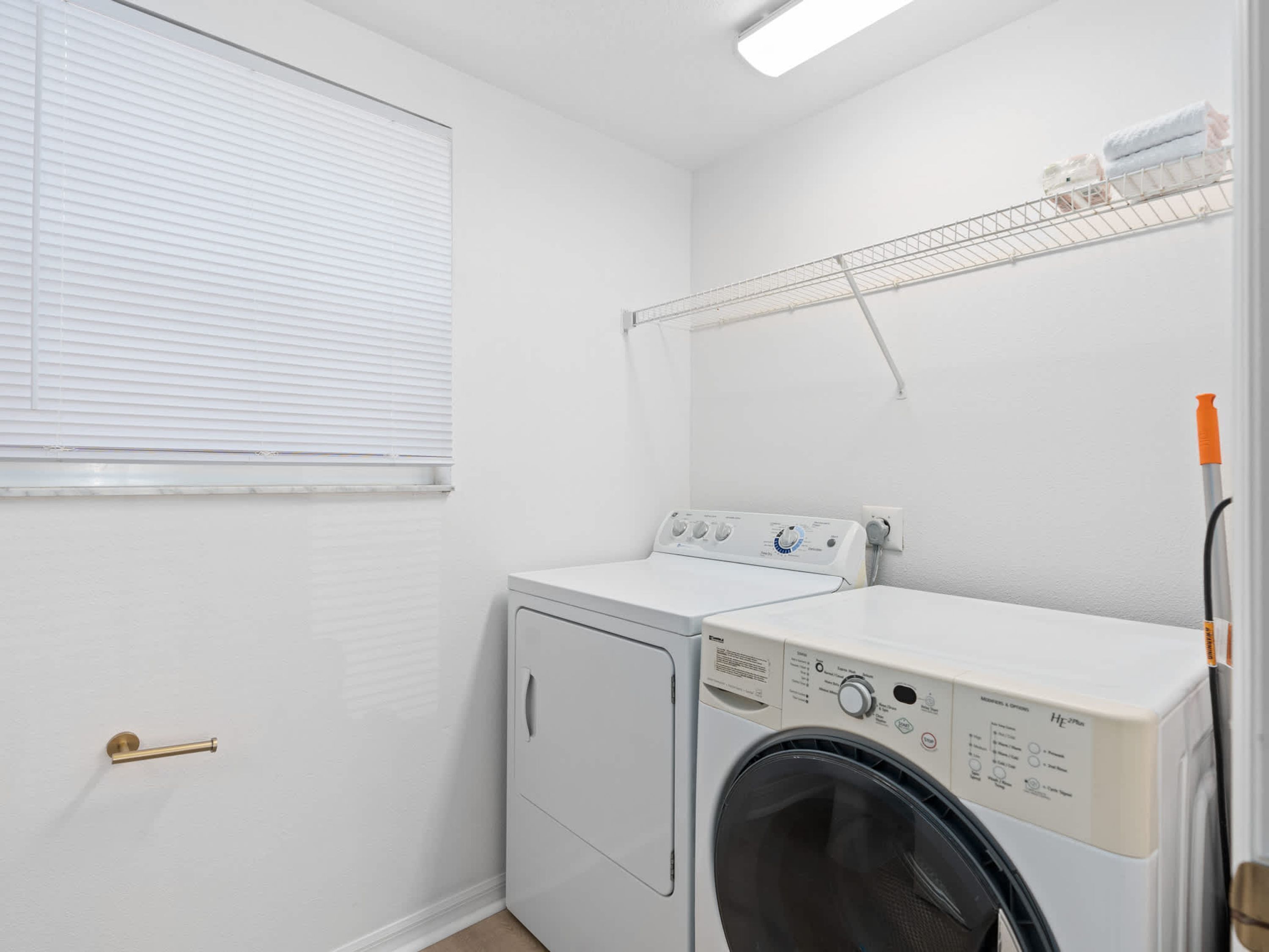 A laundry room with a front-loading washer and dryer, a metal shelf above the dryer, and a window with blinds.