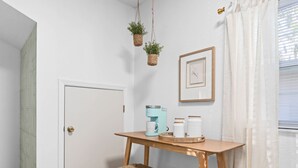 Ein kleiner, sauberer Raum mit einer weißen Tür, einem Holztisch, zwei Korbstühlen, einer Kaffeemaschine und einem gerahmten Bild an der Wand.