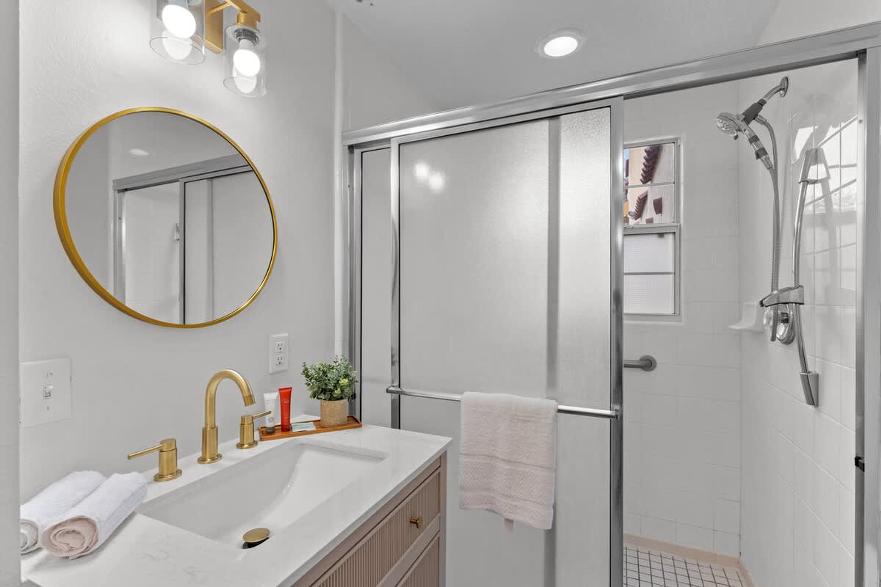 A modern bathroom with a white sink, gold faucets, a round mirror, and a glass-enclosed shower area.