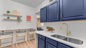 A modern kitchen with dark blue cabinets, a white countertop, and a gold faucet. There are three bar stools and a wooden cutting board on the counter. - Boho Lake Fishing Civic Center More (Kissimmee)