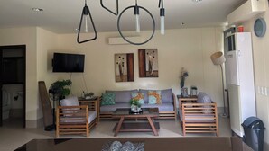 A modern living room with a sofa, coffee table, and two wooden chairs. There are ceiling lights, a flat-screen TV, and artwork on the wall.
