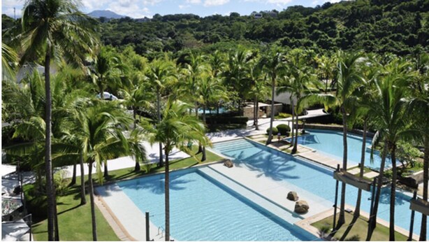 A resort pool area with multiple swimming pools, surrounded by palm trees and greenery.
