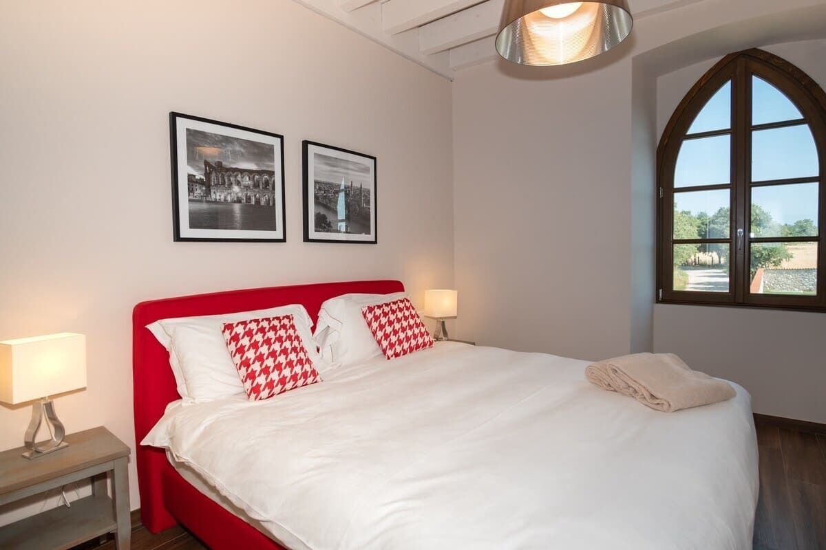 Ein Schlafzimmer mit rotem Kopfteil, weißer Bettwäsche, zwei gerahmten Bildern an der Wand, einer Nachttischlampe und einem Fenster mit Blick.