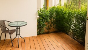 Una terraza de madera con una mesa de vidrio y una silla de metal, rodeada de abundante follaje y columnas blancas.