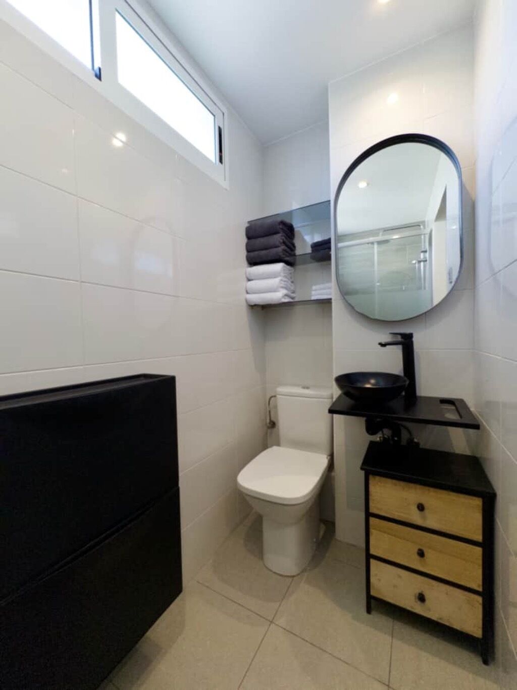 Un baño pequeño con inodoro, un lavabo de cuenca negra, un espejo y un perchero con toallas dobladas.
