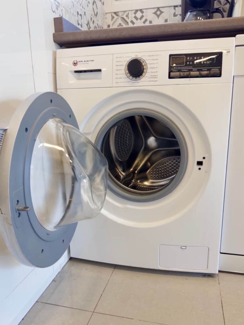 A front-loading washing machine with a transparent door, control panel, and a small storage compartment below the door.