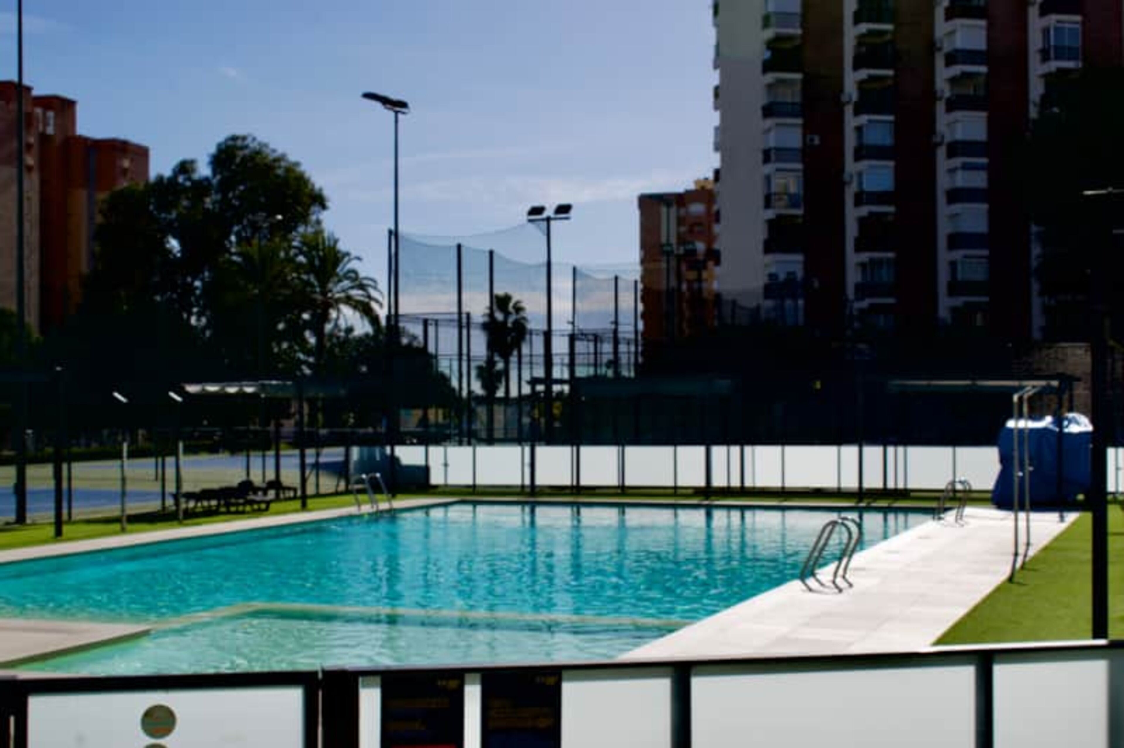 Une piscine entourée d’une clôture, bordée d’immeubles et d’arbres.