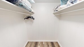 Ein kleiner, sauberer Raum mit weißen Wänden und Holzboden. Links an der Wand befinden sich zwei weiße Regale mit gefalteter Kleidung und ein schwarzer Kleiderbügel.